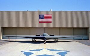 Front view of tailless aircraft parked in front of building. On the building face is a blue and red rectangular flag. A star-shaped artwork is on the taxiway in front of aircraft.